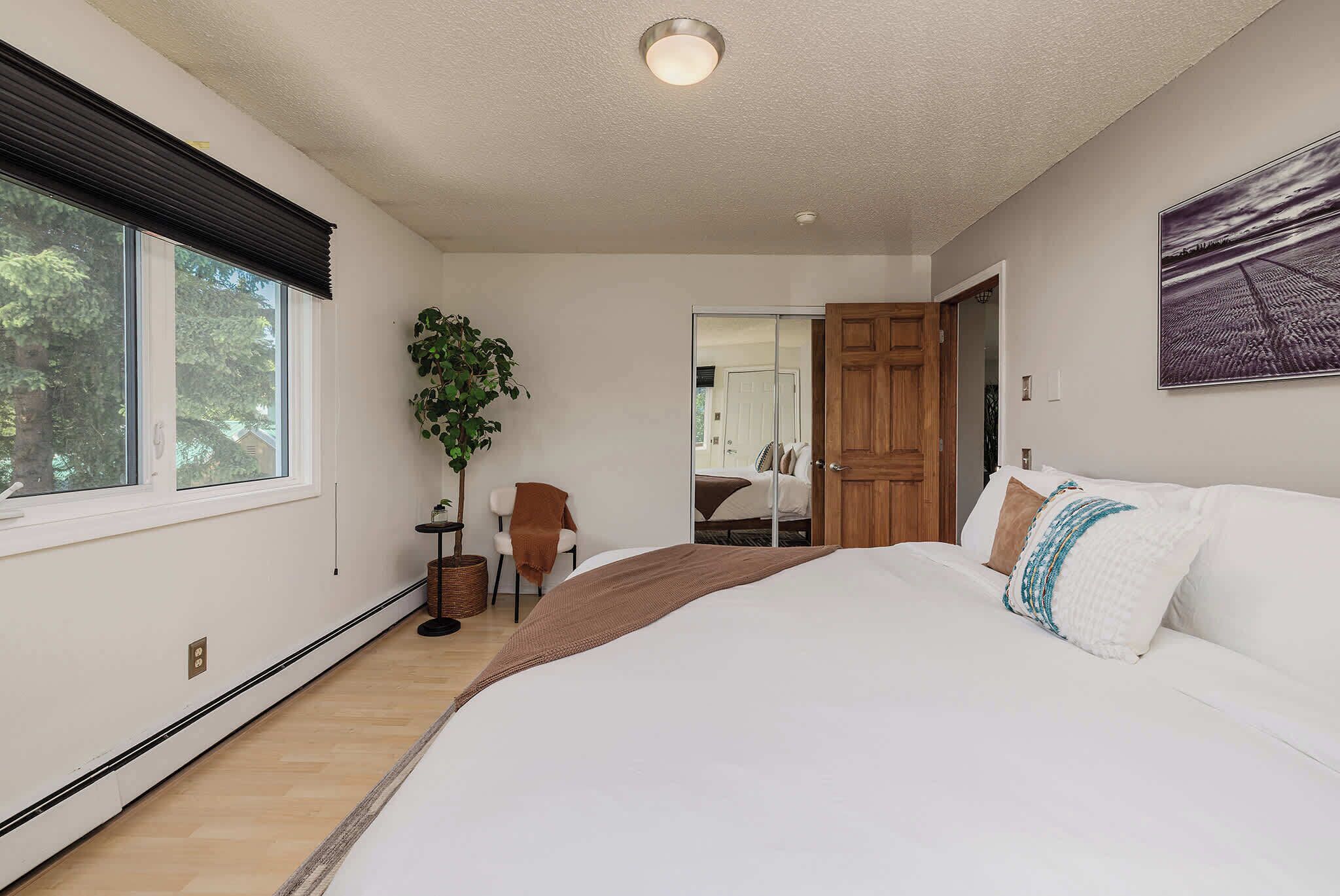 A bedroom with a large bed, a chair, a plant, and a door leading to another room.