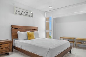 A bedroom with a wooden bed, white bedding, a yellow pillow, a nightstand, a wooden side table, and a window with a view. - Isla Verde Beachfront ESJ Tower by Otium (Carolina)