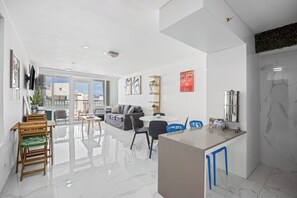 A modern living room with a sofa, dining table, and chairs, a kitchen area with a marble backsplash, and a balcony with a view. - Isla Verde Beachfront ESJ Tower by Otium (Carolina)