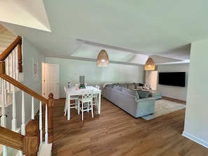 A spacious living area with a wooden staircase, a dining table with chairs, a grey sofa, and a mounted TV. - Family Retreat-Reynolds-Sleeps 12-Bunk Room-Lake Oconee Southern Charm Retreat (Greensboro)