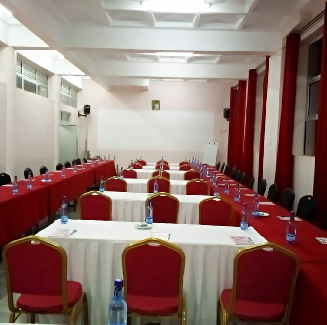 Un salón de conferencias con filas de mesas, sillas rojas y botellas de agua.