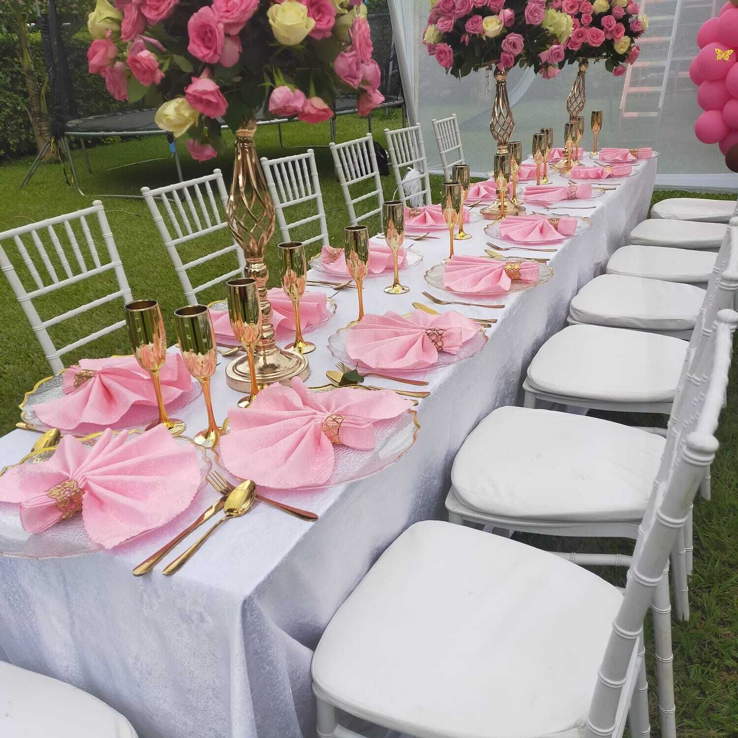 A long table set with pink napkins, gold cutlery, and clear glassware, surrounded by white chairs.
