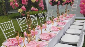 Una larga mesa vestida con servilletas rosas, cubiertos dorados y copas de vidrio transparente, rodeada de sillas blancas.