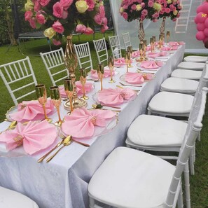 Una larga mesa vestida con servilletas rosas, cubiertos dorados y copas de vidrio transparente, rodeada de sillas blancas.