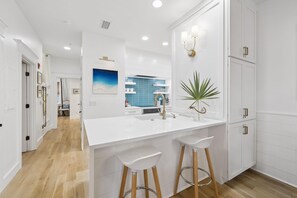 A modern kitchen with white cabinetry, a central island with stools, and a view into the dining area. - PRIVATE BEACH/POOL ACCESS-Newly Renovated-Charming Coastal Getaway (Rosemary Beach)