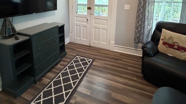 Un salon doté d’un téléviseur à écran plat, d’un canapé de couleur sombre, d’un tapis à motifs et d’une porte à deux battants donnant sur un espace verdoyant.