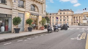 A street with a building labeled 'Mouillé' on the left, potted plants on the sidewalk, and a scooter parked on the street. - EXPERIENCE PARISIAN ELEGANCE, LOCATED IN BETWEEN INVALIDES & SAINT-GERMAIN (Paris)