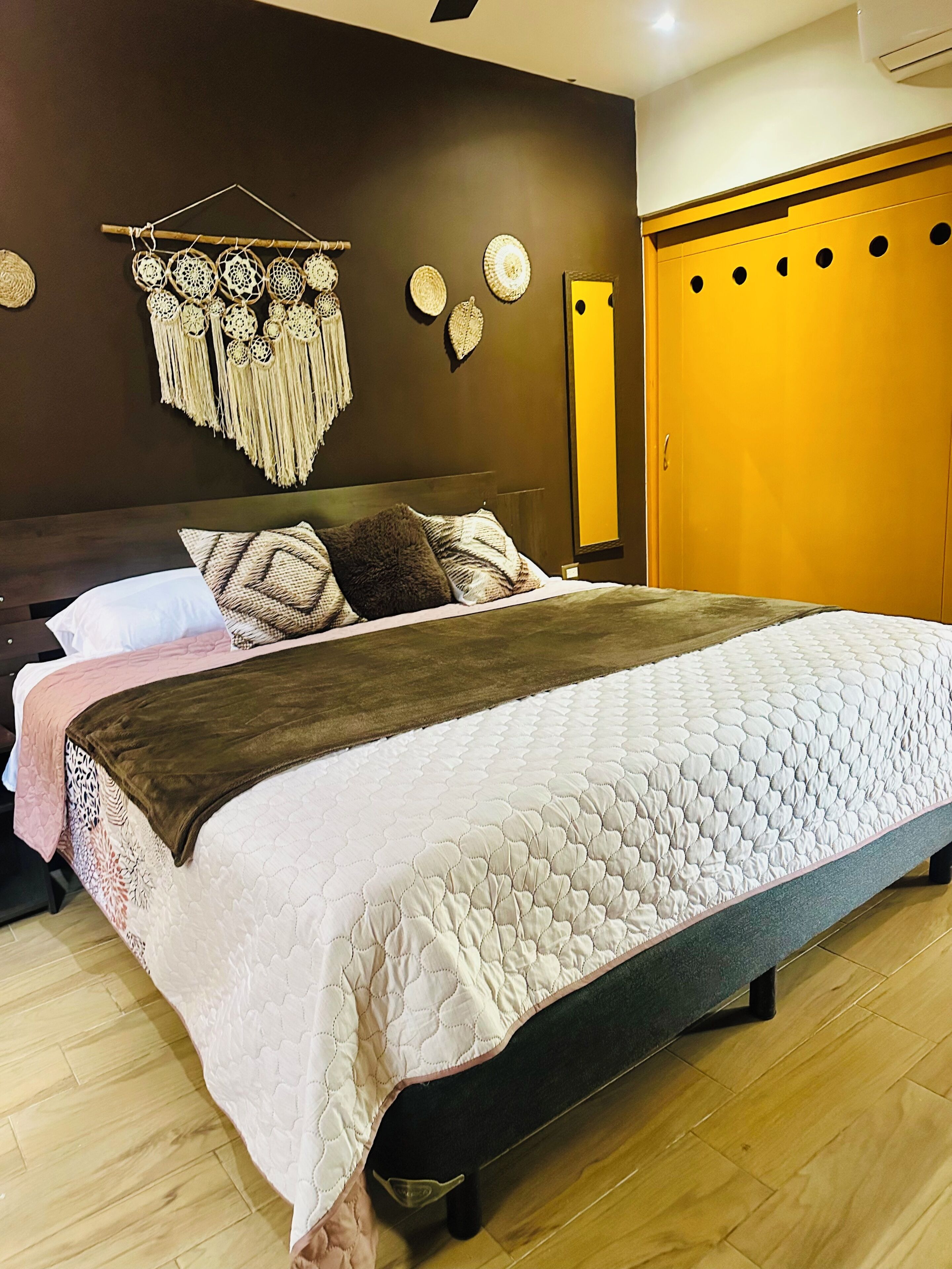 A neatly made bed with a quilted cover, a wooden headboard, and a wall-mounted macrame decoration.