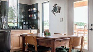 A modern kitchen with wooden cabinets, a dining table with chairs, and a large window. - Wylde Skyes | Low-Tox Retreat + Hot Tub (Fredericksburg)