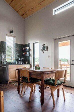 A modern kitchen with wooden cabinets, a dining table with chairs, and a large window. - Wylde Skyes | Low-Tox Retreat + Hot Tub (Fredericksburg)