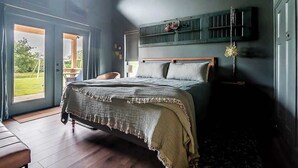 A bedroom with a large bed, a bench, a window, and a wall-mounted shelf. - Wylde Skyes | Low-Tox Retreat + Hot Tub (Fredericksburg)