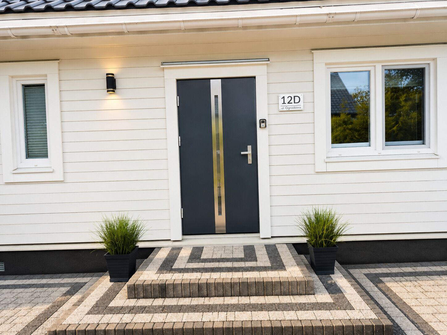 Un bâtiment blanc avec une porte sombre, deux plantes en pot et une enseigne portant le numéro « 12D ».