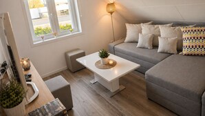 A modern living room with a sofa, a coffee table, a lamp, and a window with a view of the outside. - Willa Nordica 3 Bedroom House Garden (Stegna)