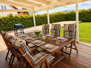 An outdoor dining area with a wooden deck, cushioned chairs, and a grill. - Willa Nordica 3 Bedroom House Garden (Stegna)