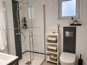 A modern bathroom with a glass-enclosed shower, white tiles, a white toilet, and a shelf with toiletries. - Willa Nordica 3 Bedroom House Garden (Stegna)