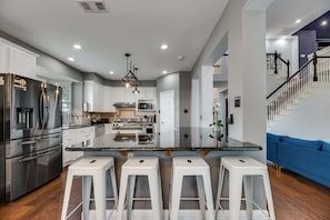 A modern kitchen with a central island, white bar stools, stainless steel appliances, and a staircase leading to a living area. - Grand Gem for Large Groups: Pool, Cinema, Playroom (Carrollton)