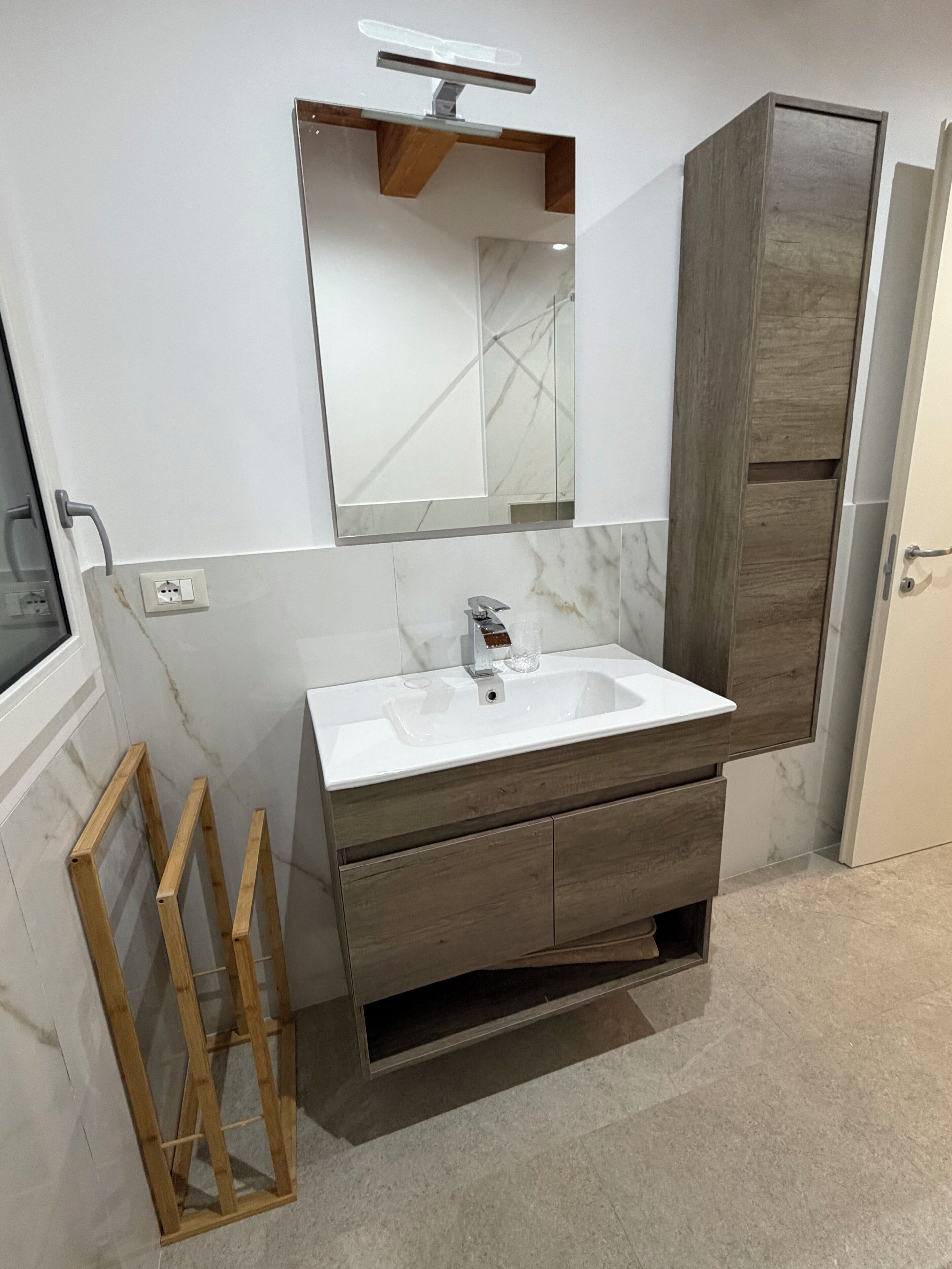 Ein Badezimmer mit einem weißen Waschbecken, einem Holzhocker, einem Spiegel und einer Tür.