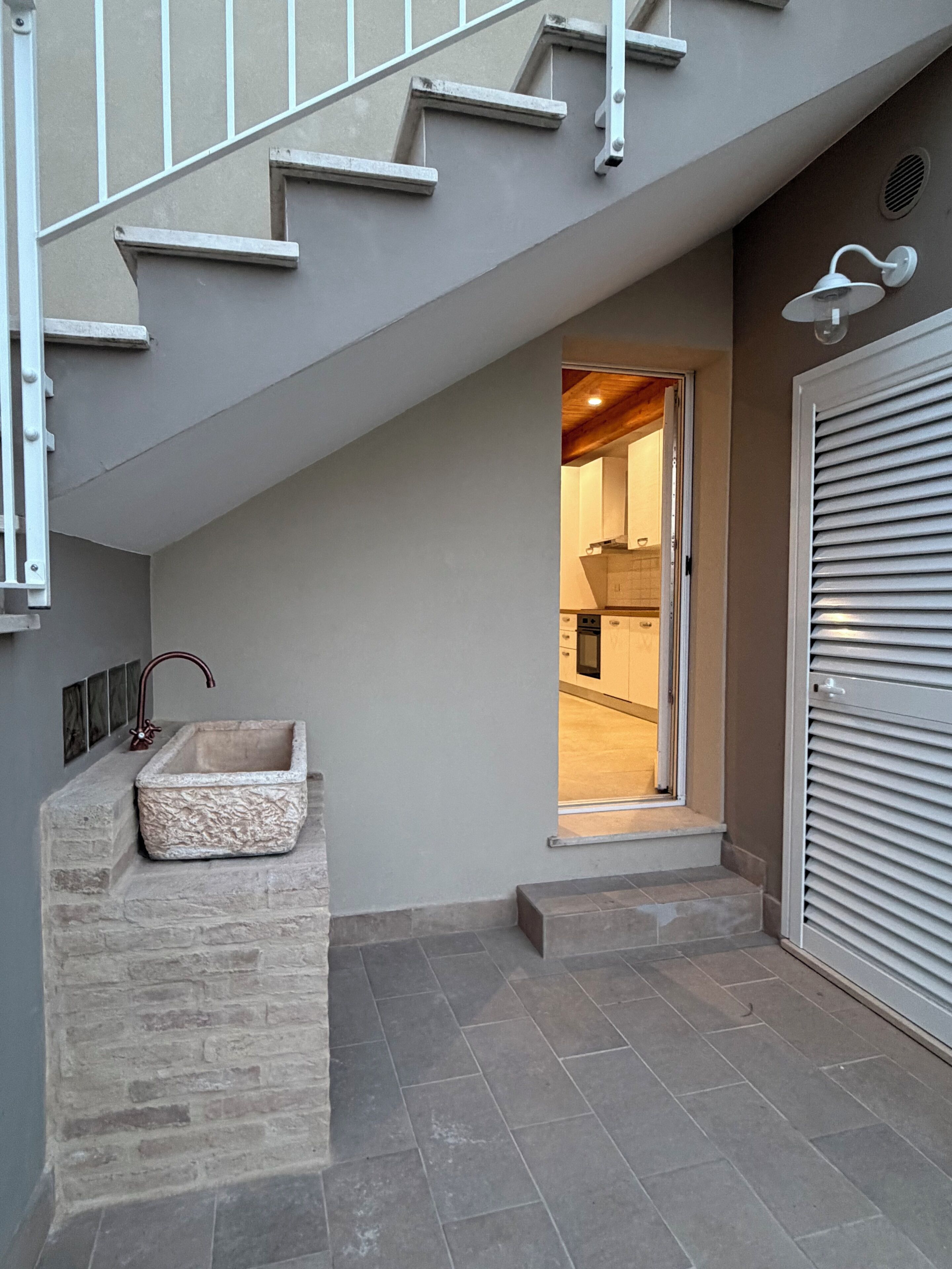 Un escalier menant à une pièce où se trouve un lavabo en pierre et une porte équipée de stores horizontaux.