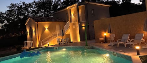 A swimming pool with a small boat, a staircase leading to a second floor, and outdoor seating with umbrellas.