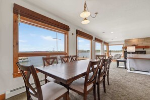 A dining area with a large table and chairs, large windows with a view of the water, and a kitchen area with a countertop and hanging light fixture.