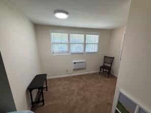 Ein Zimmer mit beigem Teppich, einem Tisch, einem Stuhl und drei Fenstern mit Jalousien.