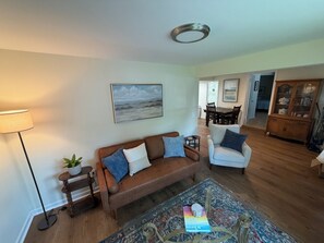 Ein Wohnzimmer mit braunem Sofa, einem weißen Sessel, einer Stehlampe, einem Gemälde und einem Essbereich im Hintergrund.