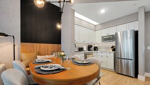 A modern kitchen with white cabinets, a stainless steel refrigerator, and a dining area with a wooden table and grey chairs. - 1 bedroom apartment near Disney and Universal (Orlando)