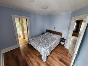 A bedroom with a wooden floor, a bed with a patterned bedspread, a nightstand, and a door leading to another room. - Cozy and Sunny 1-bedroom Apartment in Holyoke (Holyoke)
