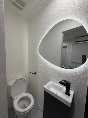 A bathroom with a white toilet, a sink with a black faucet, and a mirror with a textured wall. - The Hideaway Cabin 246 (Oistins)