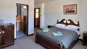 Habitación de hotel con cama, cómoda de madera, baño visible a través de una puerta abierta y un cuadro en la pared.