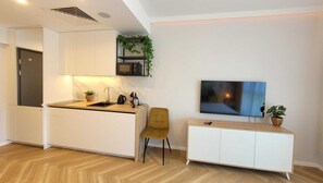 Cocina moderna con armarios blancos, encimera de madera y microondas integrado. Hay una planta pequeña en la pared y un televisor montado sobre un armario blanco.