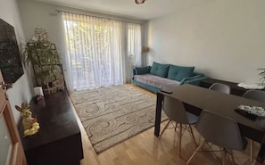 A living room with a sofa, a dining table, chairs, a rug, a plant, and a window with curtains.