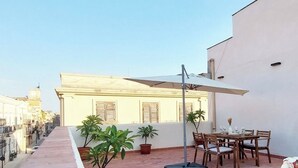 A rooftop terrace with potted plants, a dining table with chairs, and a white building in the background.