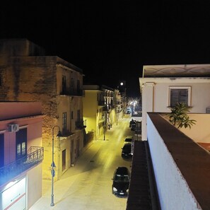 Una calle estrecha de noche con edificios a ambos lados, un coche aparcado a la derecha y una torre del reloj a lo lejos.