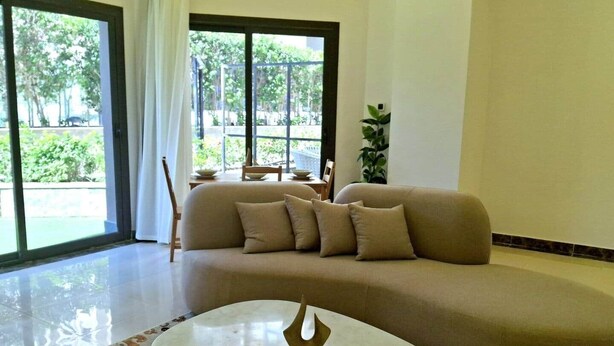 A modern living room with a beige sofa, a marble coffee table, and a view of a garden through large windows.