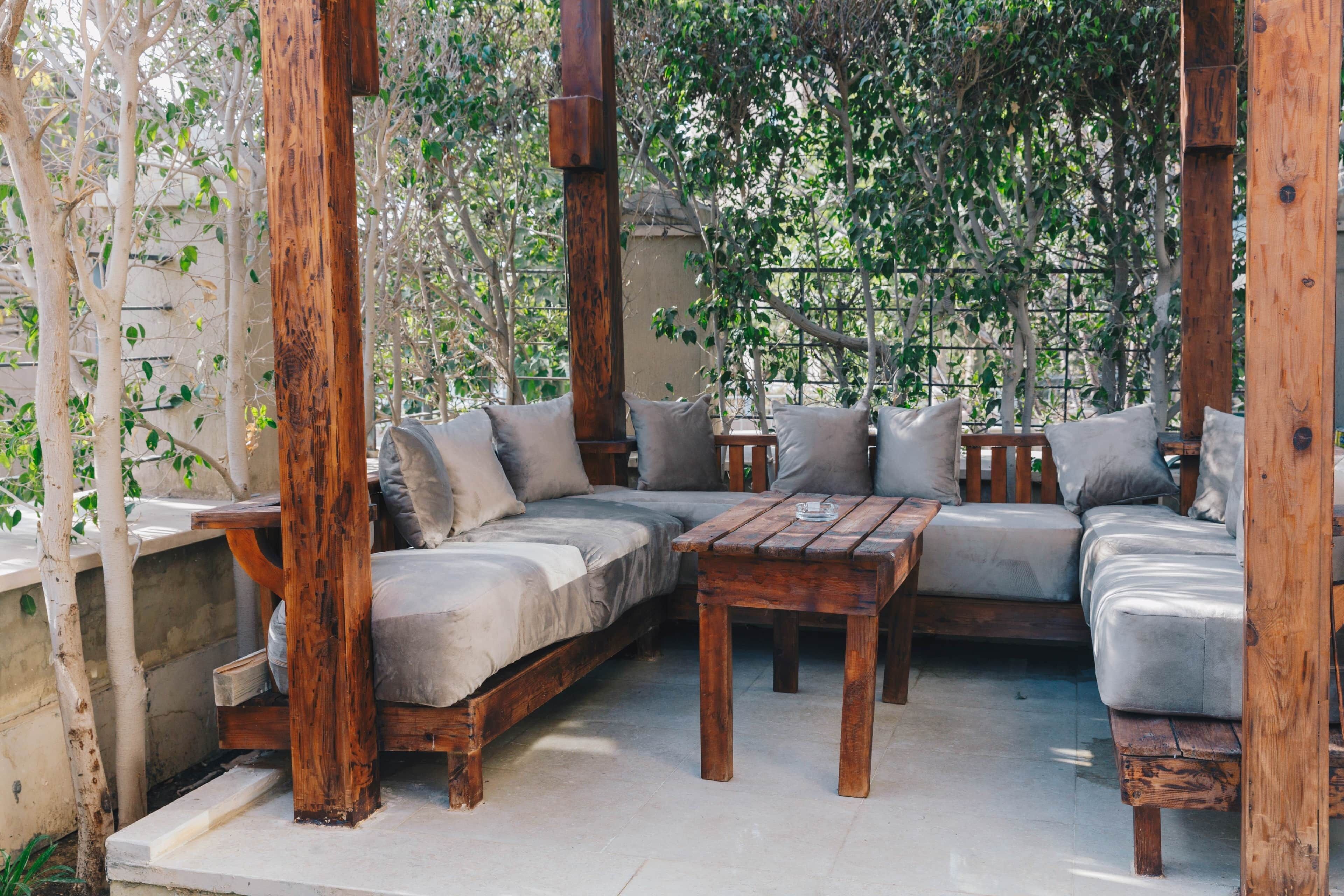 Un espace extérieur aménagé avec un banc en bois et un canapé, entouré d’arbres et d’une clôture.
