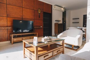 A modern living room with a flat-screen TV, wooden furniture, a glass coffee table, and a white armchair. - The Suite, Palm Hills - V702 (New Cairo City)