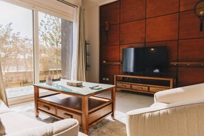 A living room with a glass coffee table, a sofa, a TV on a wooden stand, and a sliding glass door leading to an outdoor area. - The Suite, Palm Hills - V702 (New Cairo City)