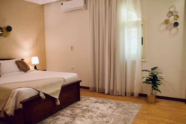 A bedroom with a bed, bedside lamp, air conditioning unit, window with curtains, wall decor, and a potted plant. - Maple and Moon Suite - Comfort & Elegance Stay 5 (New Cairo City)