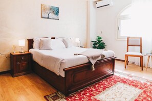 Ein ordentlich eingerichtetes Schlafzimmer mit einem hölzernen Bett, Nachttischen, einem kleinen Tisch, einem Sessel und einem großen Fenster.