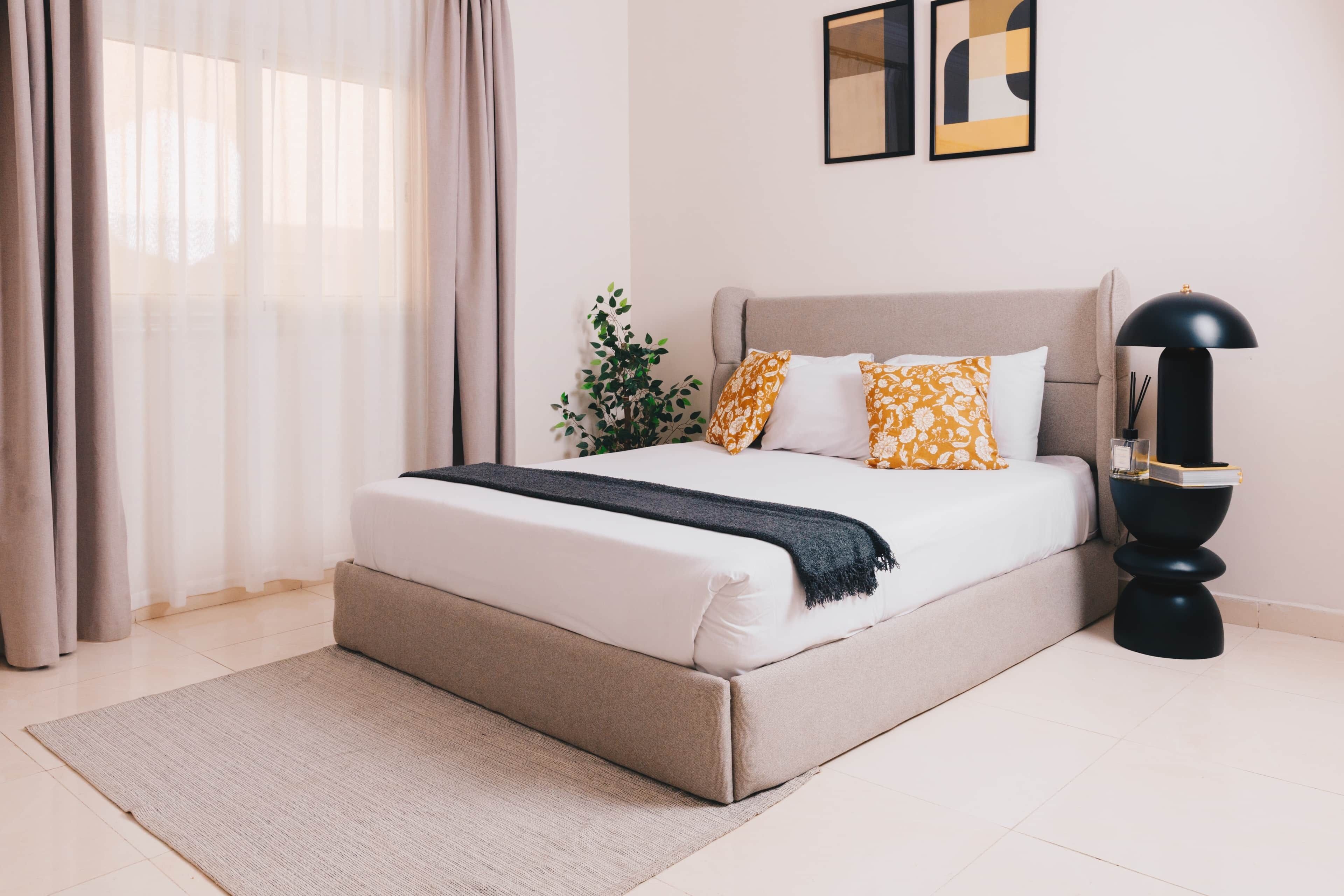 A bedroom with a bed, curtains, a plant, a lamp, and artwork on the wall.