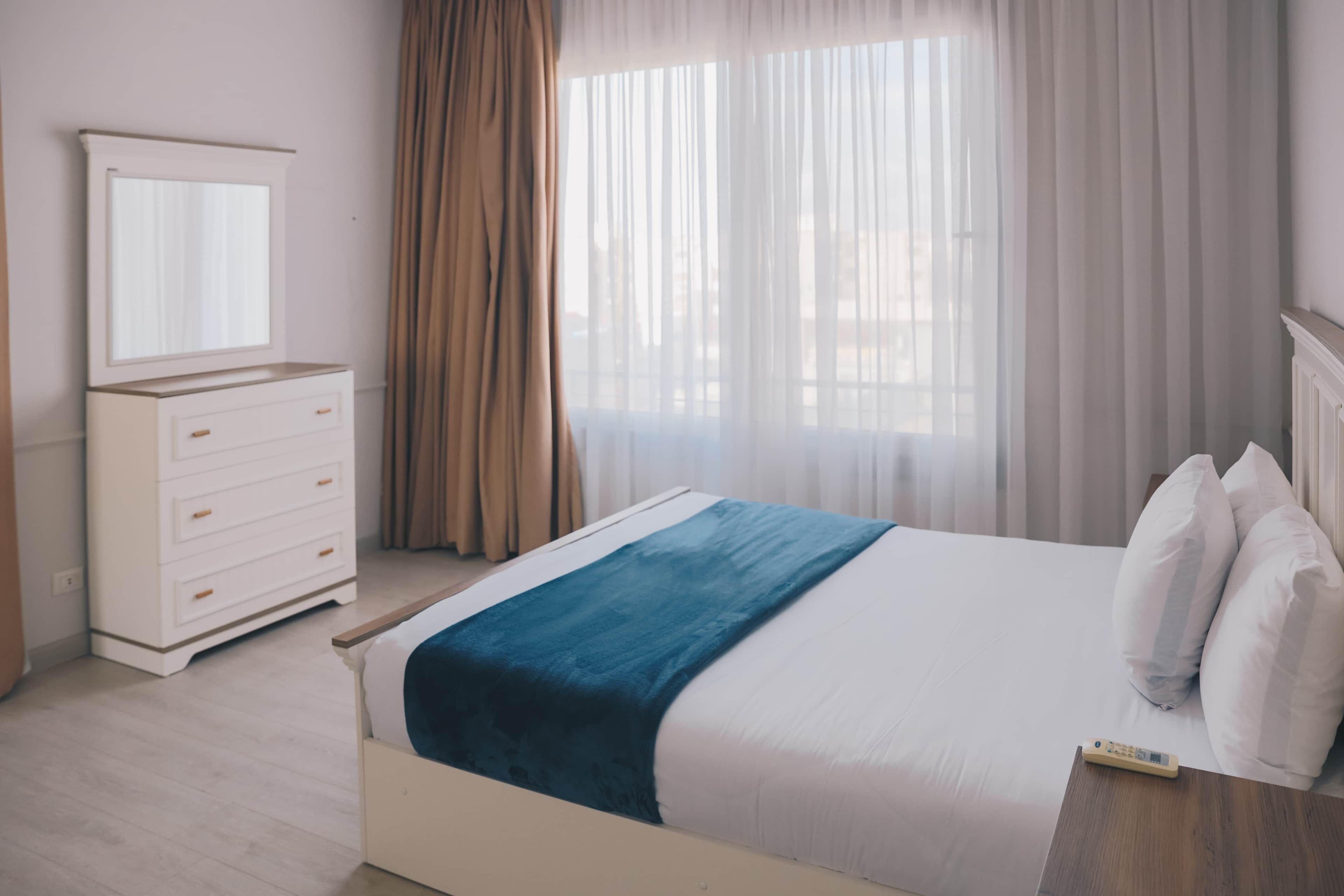 Un dormitorio con cama, cómoda, espejo y ventana con cortinas.