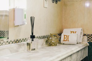 Ein Badezimmer mit Marmortischplatte, einer Handseifenflasche, einem Handtuch mit eingewebtem Monogramm und einer wandmontierten Leuchte.