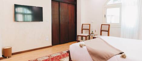 Un dormitorio con cama, televisor, armario de madera, una silla, una mesita y una ventana.