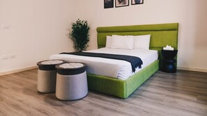 Een keurig opgemaakt slaapkamer met een bekleed bed in groene stof, wit beddengoed, twee ronde voetenbankjes, een zwart nachtkastje en een potplant.