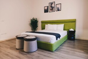 A neatly arranged bedroom with a green upholstered bed, white bedding, two round ottomans, a black side table, and a potted plant. - VIOLA Brand New Studio - 44 (New Cairo City)