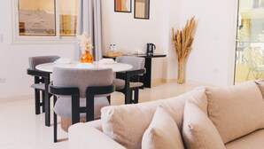 Un salon moderne avec une table à manger ronde, des chaises recouvertes d’un tissu gris, un canapé beige et un vase contenant des plantes séchées.