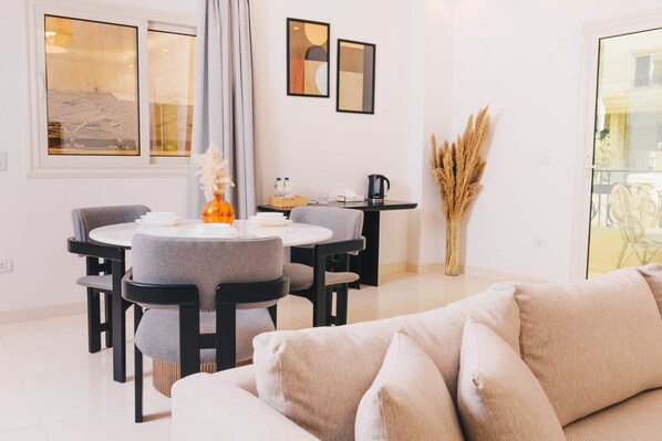 Ein modernes Wohnzimmer mit einem runden Esstisch, grau gepolsterten Stühlen, einem beigen Sofa und einer Vase mit Trockenpflanzen.