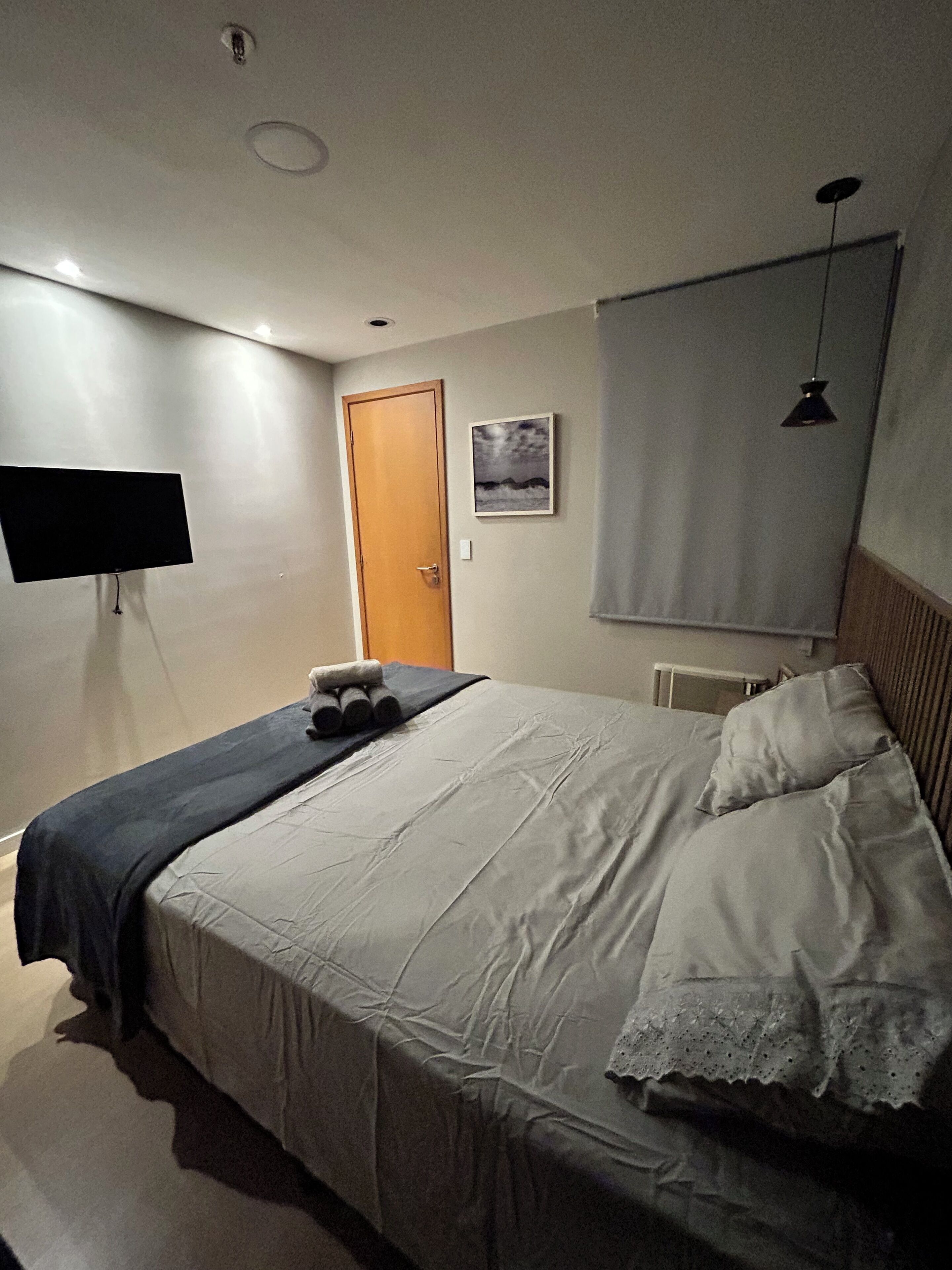 A bedroom with a bed, a flat-screen TV, a door, a picture on the wall, and a window with a blind.