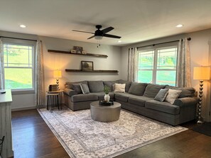 Ein Wohnzimmer mit einem Ecksofa, einem runden Couchtisch, einem Deckenventilator und großen Fenstern mit Vorhängen.